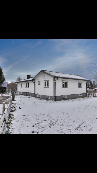 Ett tvåvåningshus med betongkällare och träpanel, snötäckt mark och ett svart staket.