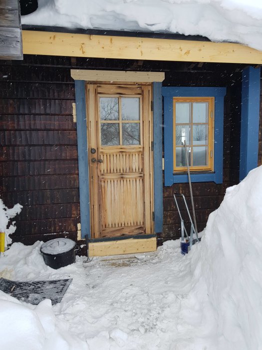 Nyinstallerad träddörr på en byggnad med snötäckt tak och omgivning, arbetsredskap syns i snön.