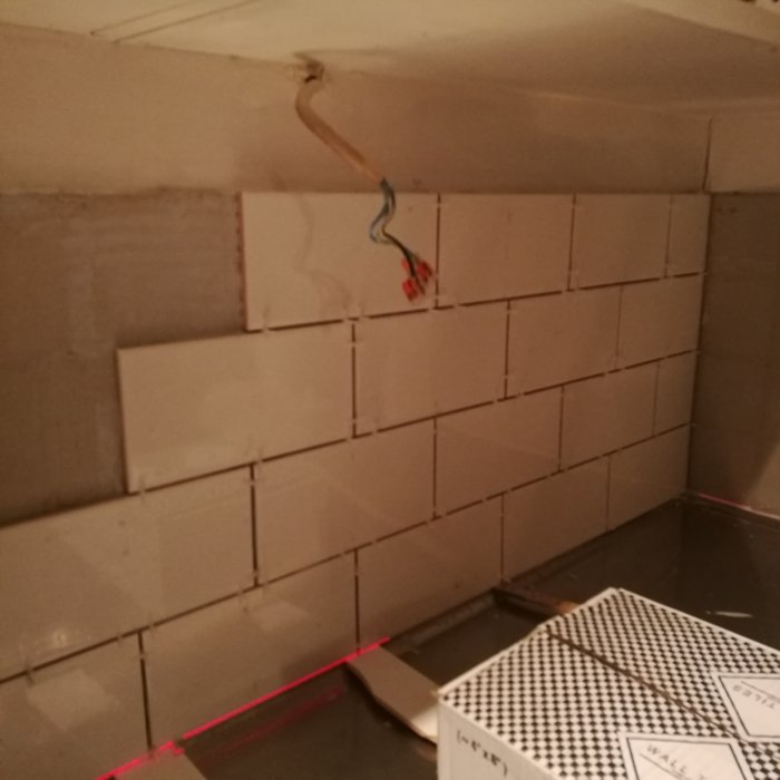 Nyinstallerade vita kakelplattor på vägg med synliga kabeländar och nivelleringslaser.