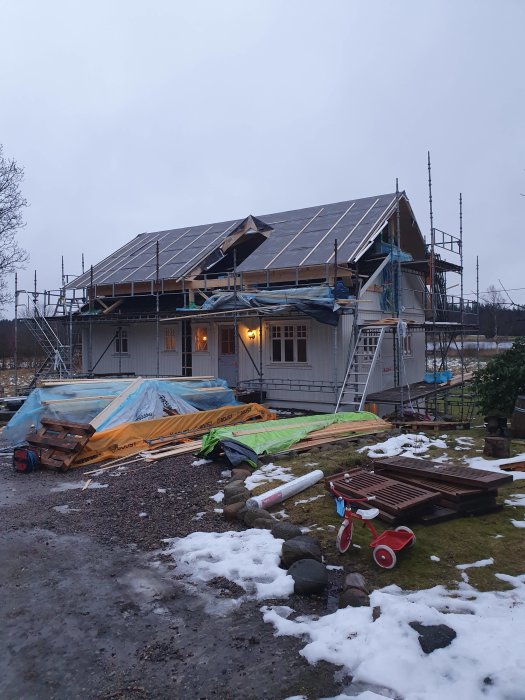 Hus under renovering med ställningar och byggmaterial spridda i trädgården, i gråväder.