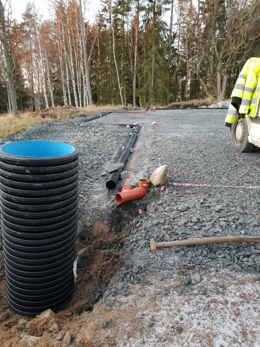 Arbetsplats utomhus med dräneringsarbeten, svart dräneringsbrunn, orangea och svarta rör samt grusunderlag med synliga måttsnören.