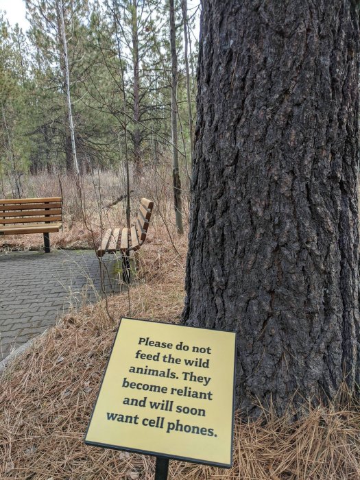 Skylt i park med text "Please do not feed the wild animals. They become reliant and will soon want cell phones." med bakgrund av träd och bänk.