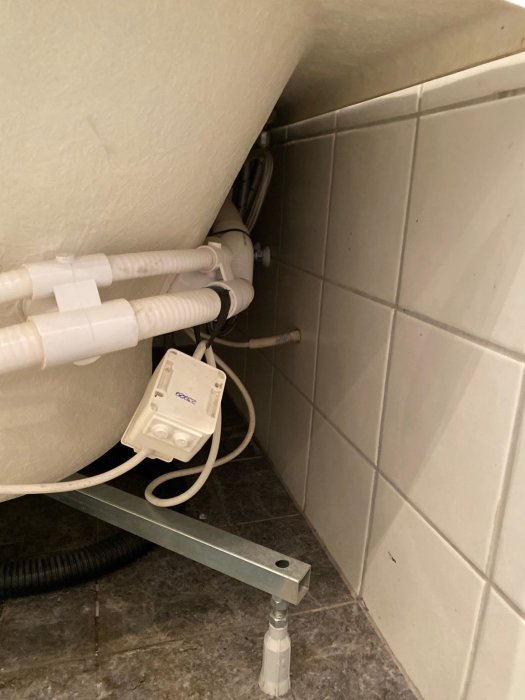 Eluttag hängande under handfat med synliga rör och kablar i ett badrum.
