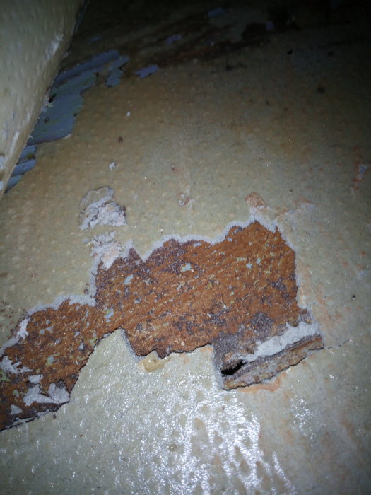 Skadat golv med brun och grå skiva som kan vara asbestpapp, flagande yta med synlig underlag.