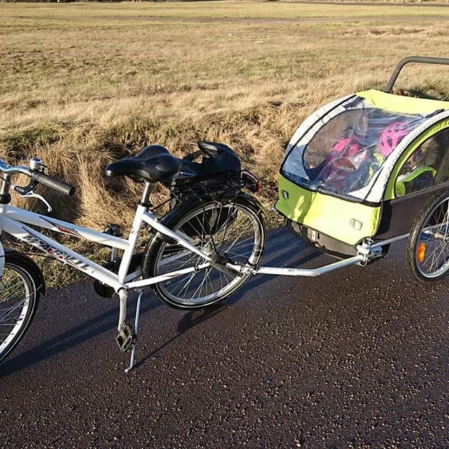 Vit cykel med barnvagnscykelkärra parkerad på en asfalterad väg bredvid ett fält.