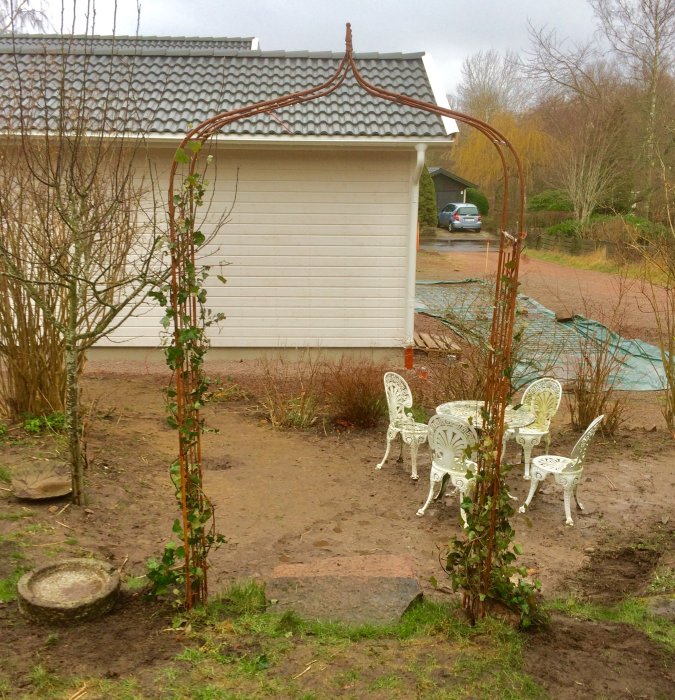 Trädgårdsställning med murgröna vid en uteplats med vita metallmöbler, intill ett hus.