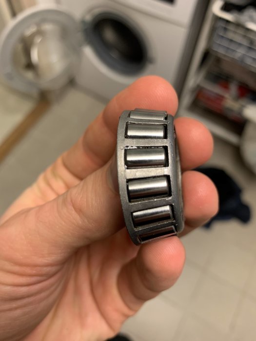 Hand håller ett renoverat kullager framför en tvättmaskin och torktumlare.