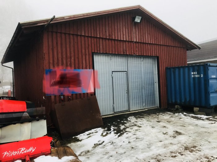 Rödfärgat garage med lockpanel, dubbla dörrar och snöfläckar i förgrunden.