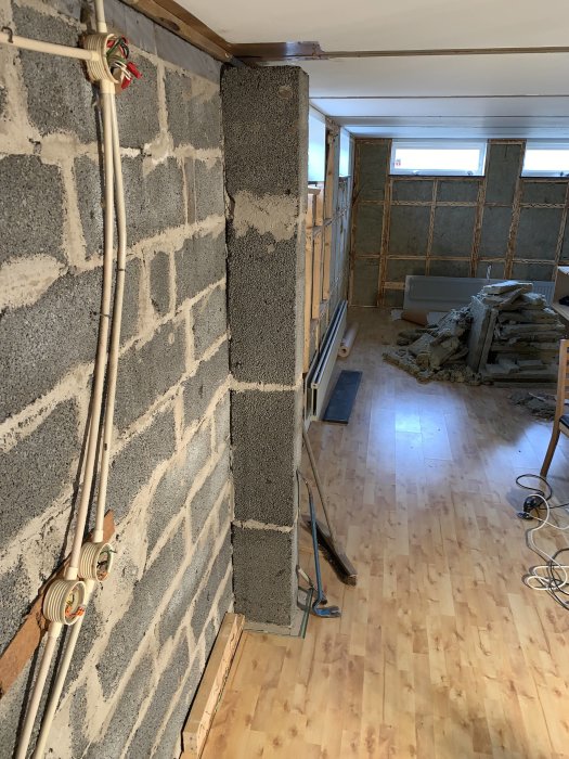 Pelare i en urtagen vägg i källare med synliga kablar och en rörböj vid golvet, omgivet av byggmaterial.