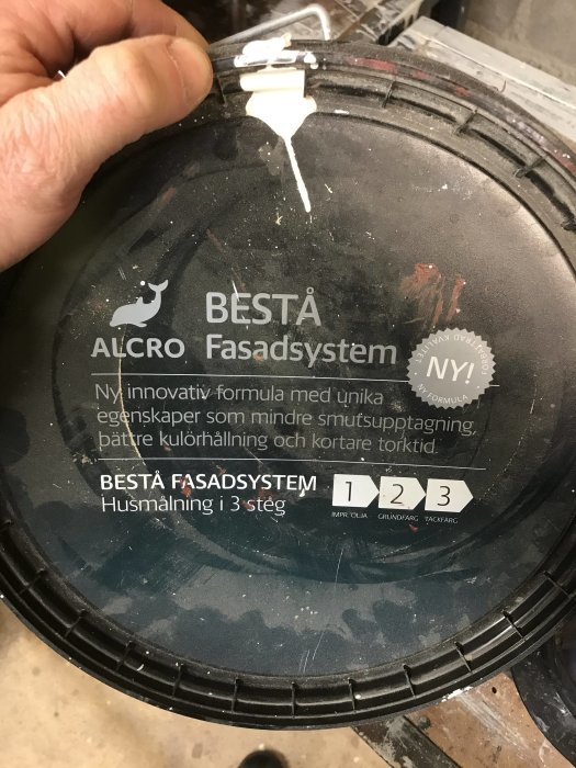 Hand håller en burk med texten 'ALCRO BÄSTA Fasadsystem' och synliga vita färgrester på locket.