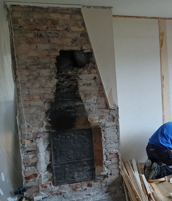Riven eldstad med murad skorsten och rester av en öppen spis, omgivet av en vit vägg och byggmaterial.