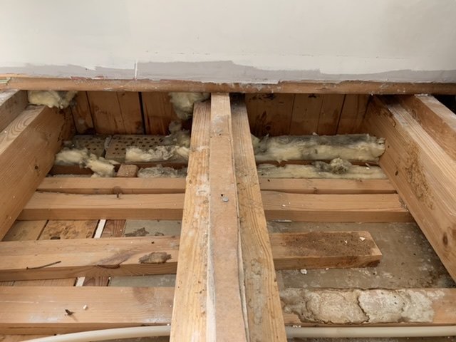 Öppnat golv i hörnrum visar bjälklag och gammal isolering utan luftspalt mot väggkonstruktion.