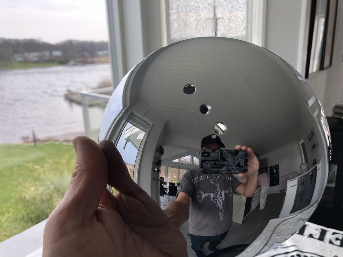 Hand håller en reflekterande lampskärm till Foscarini Caboche Picola, med kök och utsikt över sjö speglad i ytan.