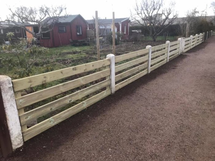 Nybyggt trästaket längs en grusgång i ett koloniområde, med röda kolonistugor i bakgrunden.