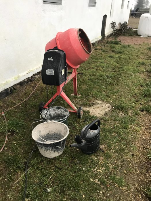 Röd betongblandare med hinkar och vattenkanna framför en vit vägg, använd vid byggprojektet beskrivet i foruminlägget.