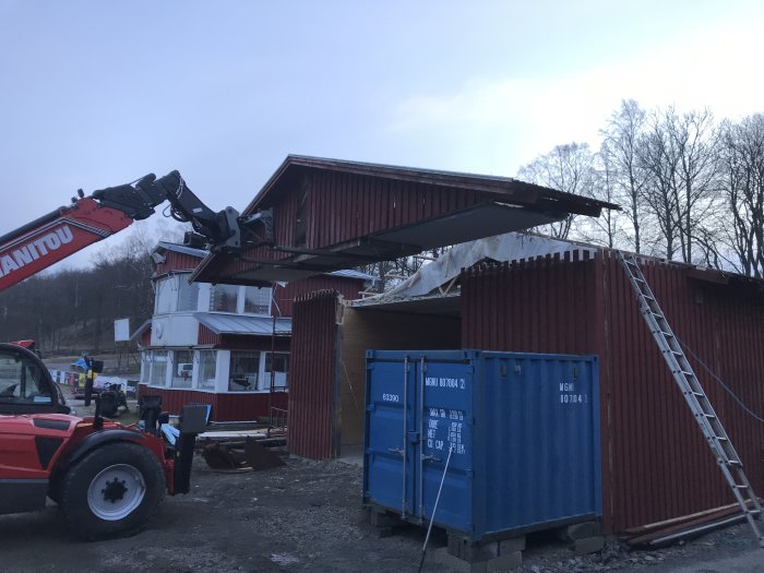 Teleskoplastare som lyfter bort plåttak från byggnad under rivningsarbete.