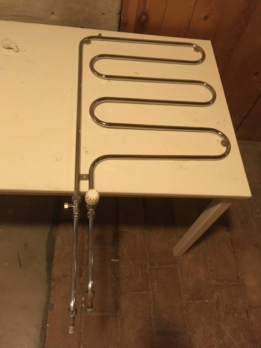 Begagnad handdukstork i metall liggande på en vit yta med anslutningsrör nedåt, troligen rostfritt stål.