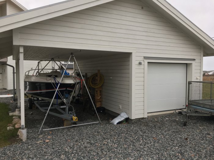 Carport ansluten till garage med makadamgolv och parkerad båt på trailer.