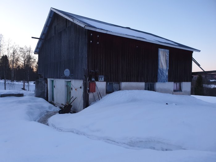 En gammal ladugårdsbyggnad med sliten fasad, täckt i snö, under en vintertidig kväll.