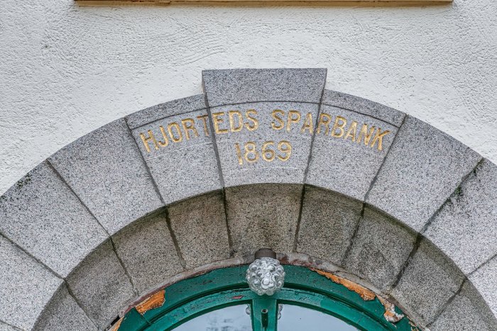 Stenvalv över dörr med texten "HJORTEDS SPARBANK 1869" på en vitmålad vägg, historisk byggnad.