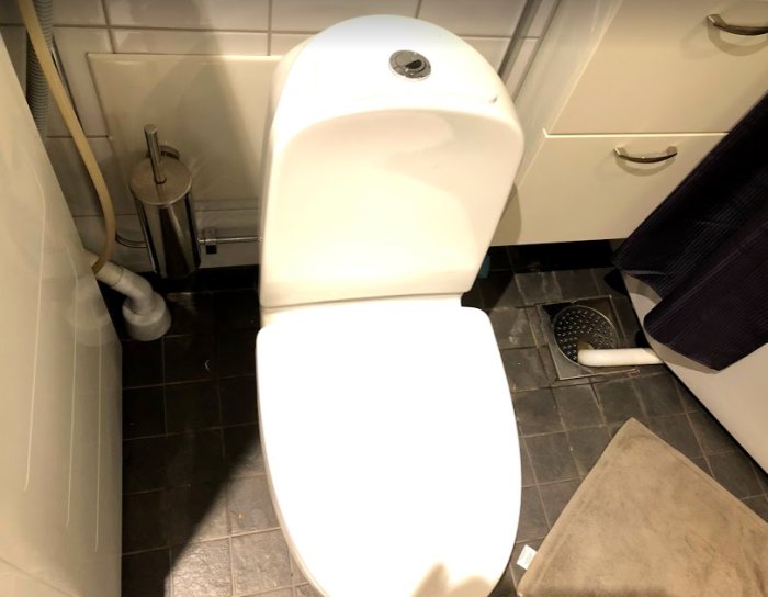 Vy av ett badrum med toalett, synliga rör och avlopp bakom inspektionslucka, ingen tätskikt synlig.