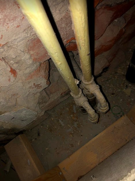 Bild på gamla tegelväggar och synliga rörinstallationer bakom en inspektionslucka i ett badrum.