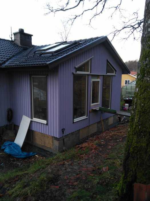 Utbyggnad av ett hus med lila fasad och ett takfönster, synliga grundarbeten och begagnade fönster.