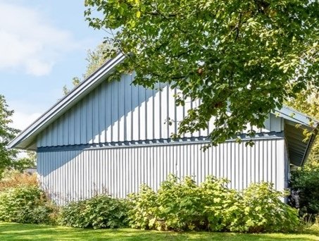Hörnet av ett hus med stående vit grundmålad träpanel, grönskande omgivning, förfrågan om renoveringskostnad.