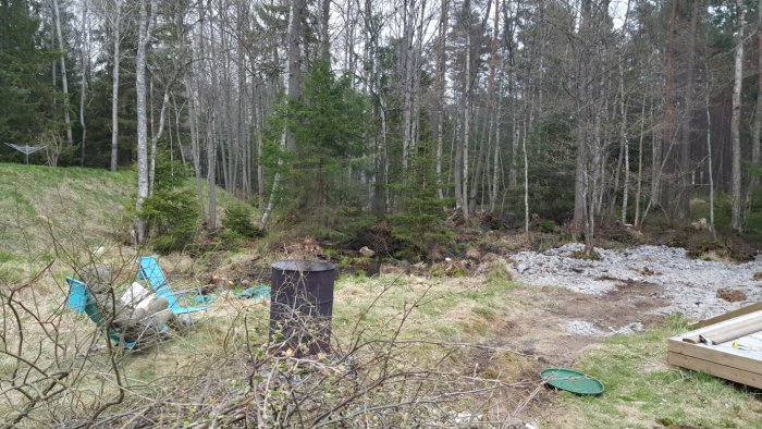 Damm i skogsmiljö med lätt vattenflöde, omgiven av träd och gräs, två blå stolar och brunnlock i förgrunden.