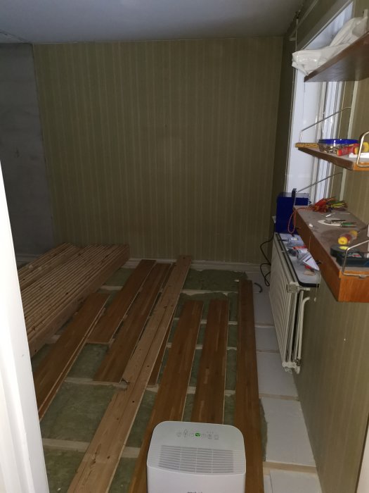 Avfuktare igång i ett rum under renovering med träreglar utlagda på golvet och tapetserade väggar.