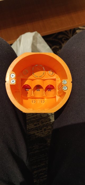 Orange installationsdosa av märket Elko med synliga låsskruvar bredvid montageskruvarna i en persons knä.