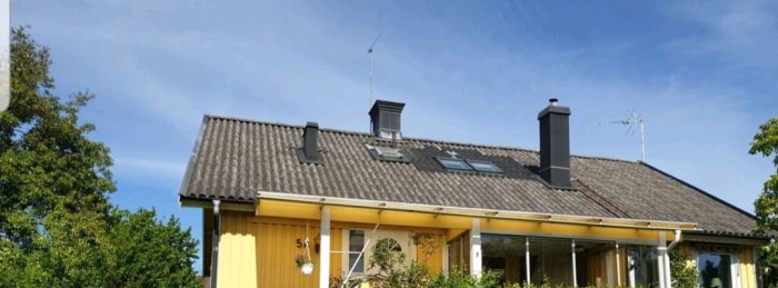 Ett hus med en mossfri takyta, skorstenar, fönster och en antenn under blå himmel.