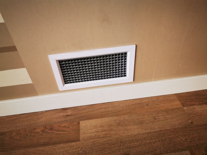 Ventilationsgaller i vägg som blåser ut luft istället för att ta in, mot trägolv och beige vägg.