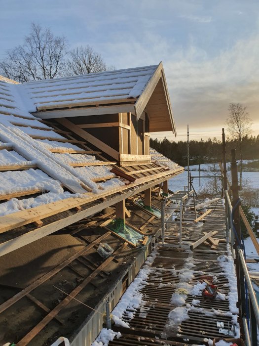 Hustak under konstruktion täckt med snö vid solnedgång, med synlig väggyta och byggställningar.