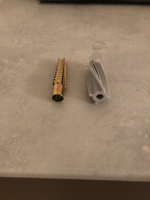 Två olika typer av lättbetongpluggar på en grå yta, en i metall och en i vit plast med vingar.