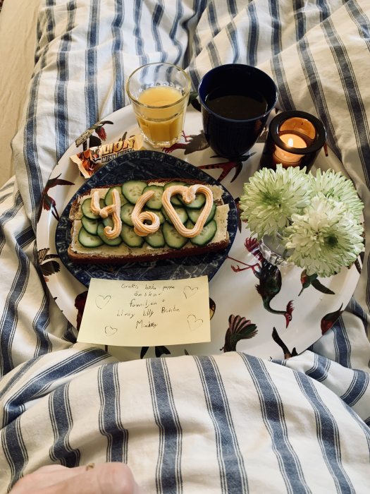 Frukostbricka på sängen med macka, juice, kaffe och en lapp med gratulationer samt blommor och ljus.