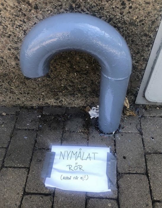 Nymålat grått rör mot en vägg med skylt "NYMÅLAT RÖR (Alltså rör ej!)" på marken.