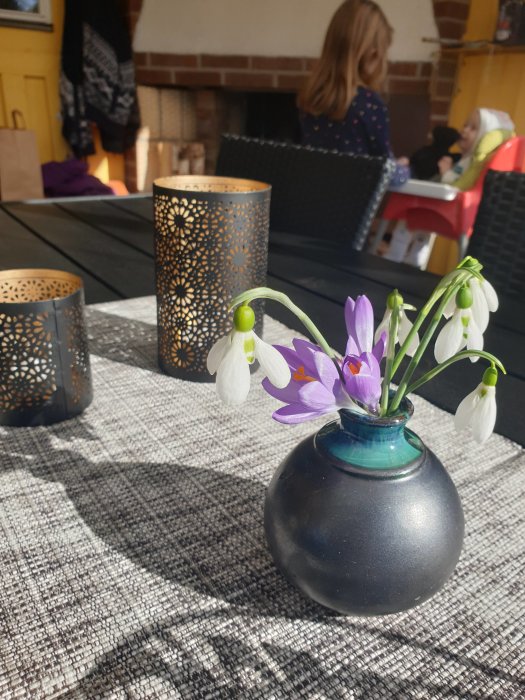 Vårblommor i vas på bord i uterum med ljuslyktor och personer i bakgrunden.