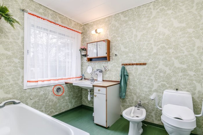 Vintage badrum med bladmönstrad tapet, badkar, handfat, och bisyssla. Rund luftventil markerad nära fönstret.