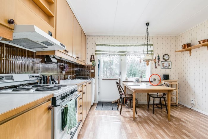 Vintagekök med träskåp, vit spis och mönstrad tapet samt cirkelformat hål vid fönstret.