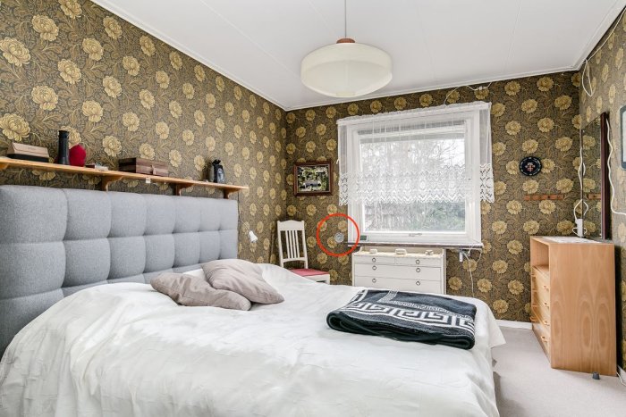 Sovrum med mönstrad tapet, säng, och cirkulärt hål nära fönster för möjlig ventilation.