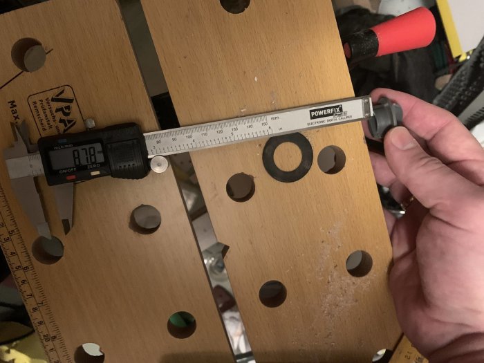 Digital skjutmått som visar 82,7 mm mot en träskiva med flera utskurna hål.