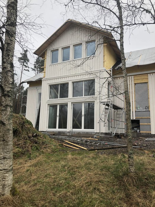 Hus under renovering med ny monterad träpanel på takkupa och fasad, stegar och byggmaterial synligt.