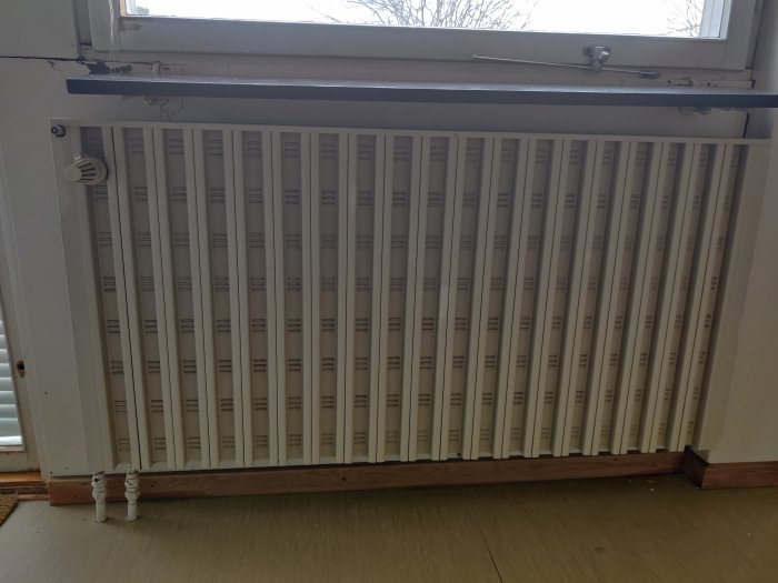 Vit radiator under ett fönster i behov av renovering med synliga rör och termostat.