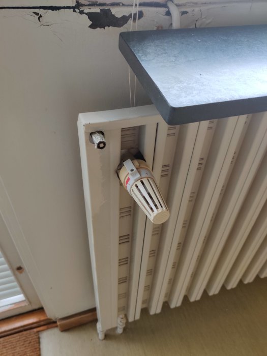 Ett vitmålat radiator-element och termostat i ett rum med skadad vägg ovanför.