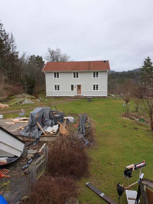 Vit panelhus med röda takpannor, omgivet av oordnat trädgårdskaos med byggmaterial och grus.