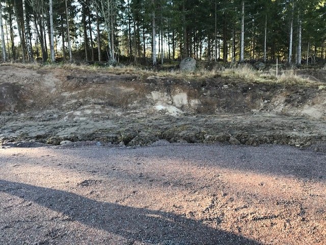 En ojämn slänt med en blandning av berg och jord vid en tomt, grus i förgrunden och träd i bakgrunden.