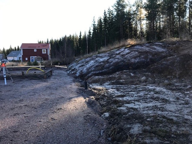 En slänt med berg och jord vid ett byggprojekt, omgiven av träd och ett rött hus i bakgrunden.