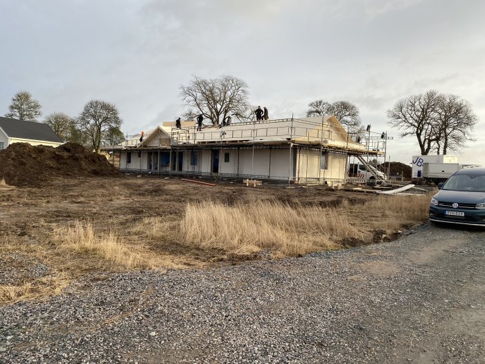 Byggarbetsplats med arbetare på ett hus under uppbyggnad, omgiven av högar med jord och byggfordon.