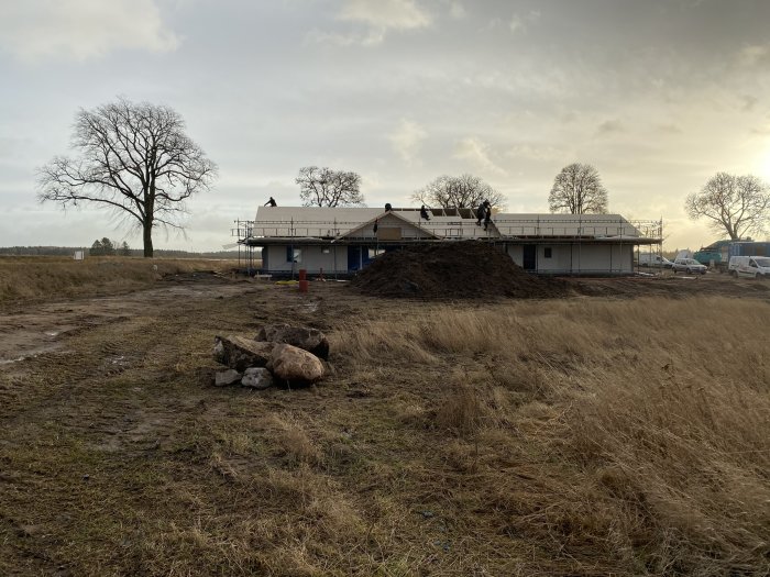 Hus under konstruktion med arbetare på taket, byggmaterial på marken, i ett landskap med träd och molnig himmel.